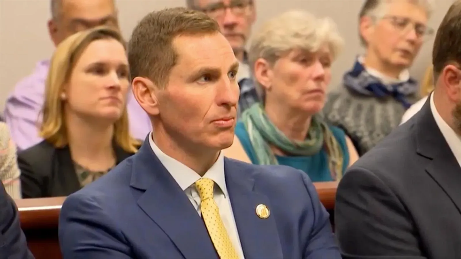 شخص يرتدي بدلة رسمية يجلس في قاعة المحكمة مع مجموعة من الأشخاص في الخلفية، يعكس أجواء الجلسات القانونية في نورث كارولينا.