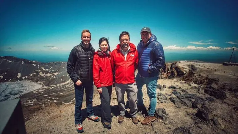 مجموعة من الصحفيين يقفون على قمة جبل بايكتو، مع منظر للبحيرة المحيطة والجبال، مستمتعين بالجو النقي والمعالم الطبيعية.