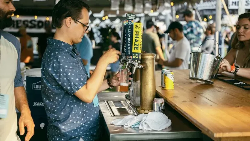 رجل يصب مشروبًا من صنبور في مهرجان محلي، مع حشود من الزوار في الخلفية، يعكس تراجع السياحة الكندية في منطقة نيويورك.