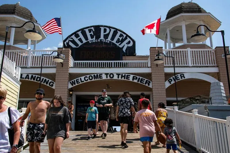 مدخل رصيف يتضمن لافتة ترحيبية، مع علم كندا وأعلام أمريكية، وزوار يتجولون في أجواء صيفية.