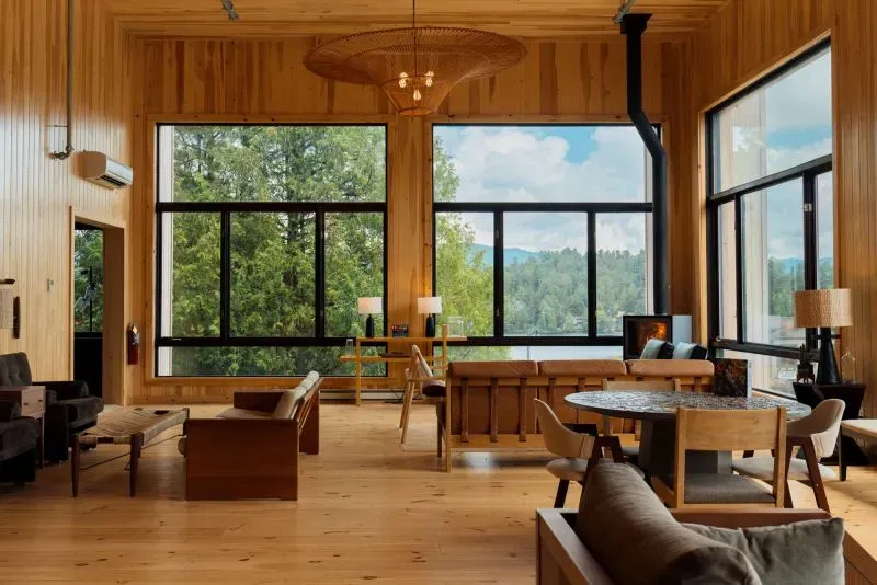 مساحة داخلية حديثة لمنزل أو منتجع، تتميز بنوافذ كبيرة تطل على المناظر الطبيعية الخلابة، مع أثاث خشبي أنيق وأجواء مريحة.