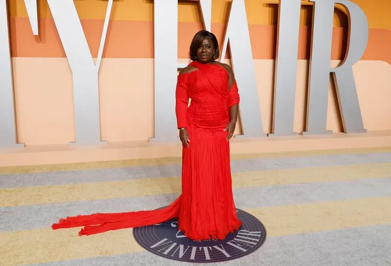 تظهر الصورة ممثلة ترتدي فستانًا أحمر لامعًا في حفل فانيتي فير، مع خلفية ملونة تعكس أجواء السهرة الراقية.