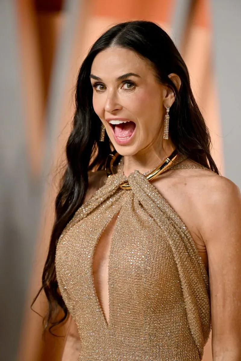 ديمي مور مبتسمة بفستان ذهبي لامع، تعكس أجواء حفلات هوليوود بعد حفل توزيع جوائز الأوسكار، وسط أضواء ساطعة واحتفالية.