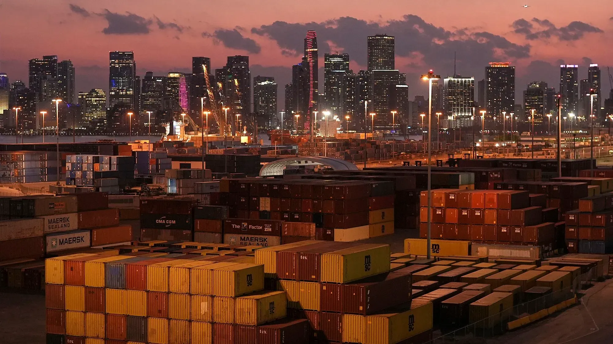 صورة لميناء مزدحم في مدينة كبيرة، حيث تكدست حاويات الشحن تحت سماء غائمة، مما يبرز تأثير التعريفات الجمركية على التجارة.