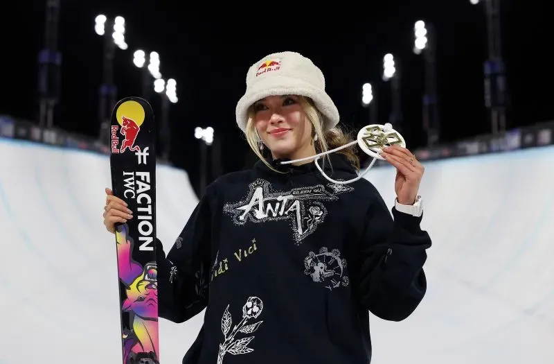 إيلين غو، المتزلجة الأمريكية، تحمل ميدالية وتبتسم بفخر، مرتدية سترة سوداء وقبعة، في موقع التزلج تحت الأضواء.