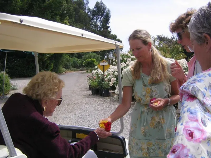 امرأة تقدم وردة ملونة لامرأة أخرى أثناء مناقشة زراعة الورود في حديقة، مع أشجار وزهور تظهر في الخلفية.