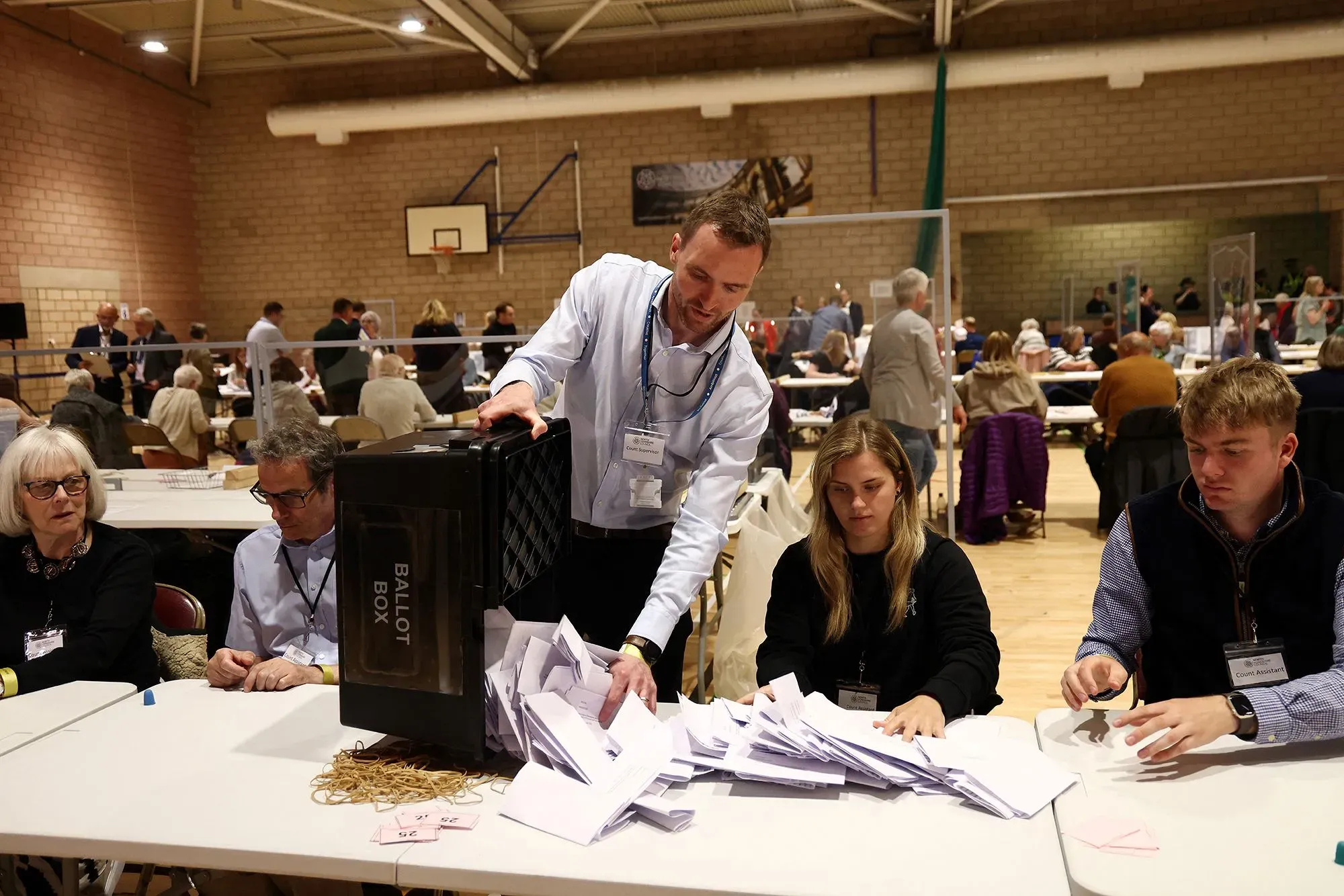 عملية فرز الأصوات في مركز اقتراع، حيث يقوم عدد من الأشخاص بتفريغ صناديق الاقتراع على طاولة، مع وجود مراقبين في الخلفية.