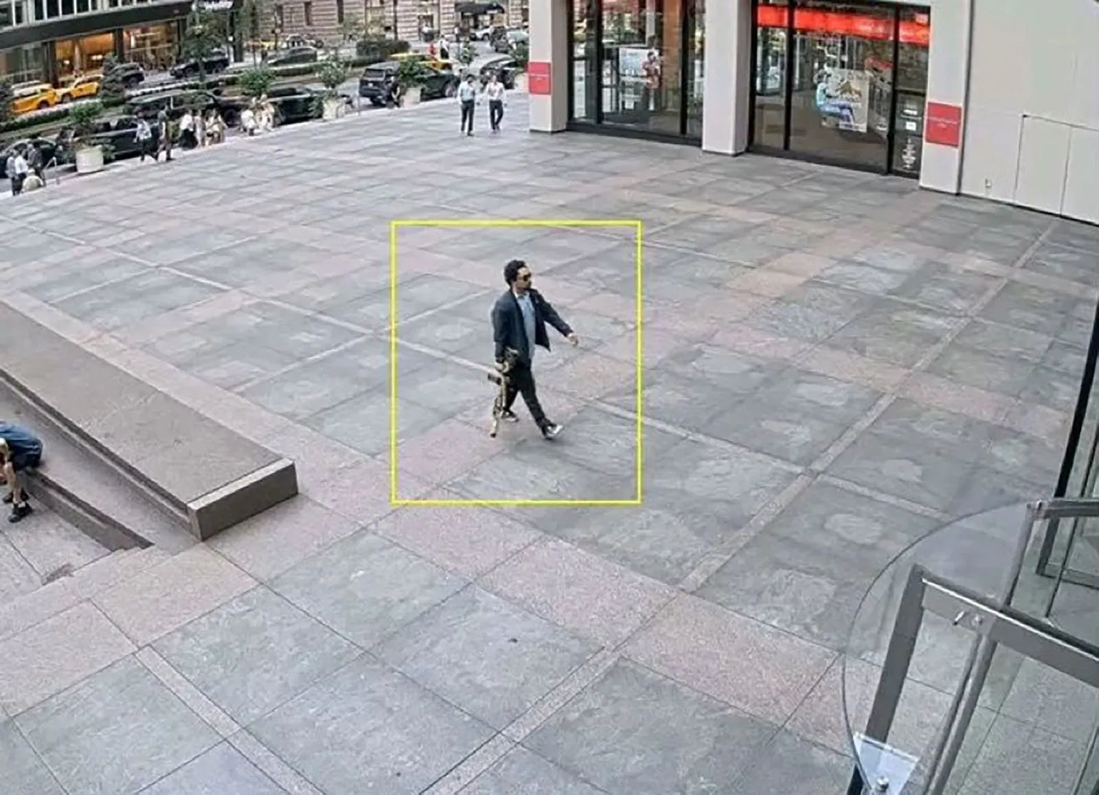 شخص يرتدي نظارات شمسية ويحمل بندقية هجومية، يسير نحو مدخل مبنى مكاتب في نيويورك، حيث وقعت حادثة إطلاق نار.