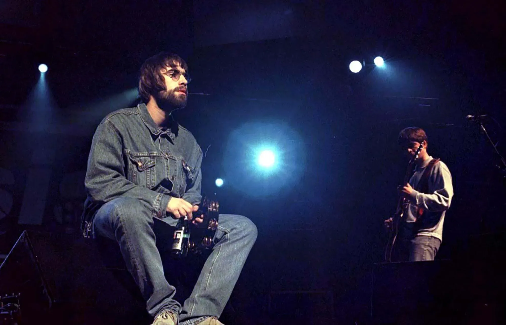 ليام غالاغر يجلس على المسرح خلال عرض لفرقة Oasis، مع إضاءة خلفية واضحة، مما يعكس توتر العلاقة بينه وبين شقيقه نويل.