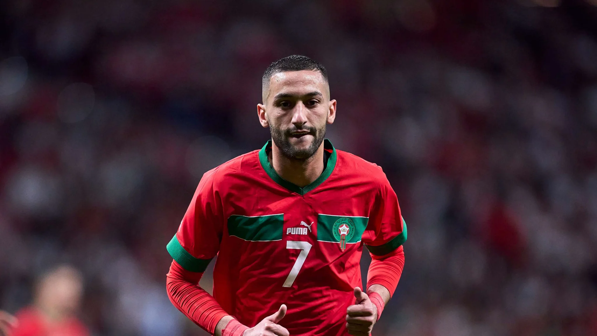 حكيم زياش لاعب المنتخب المغربي يرتدي القميص الأحمر خلال مباراة، نجم كرة القدم المغربية في لحظة تركيز عالية.