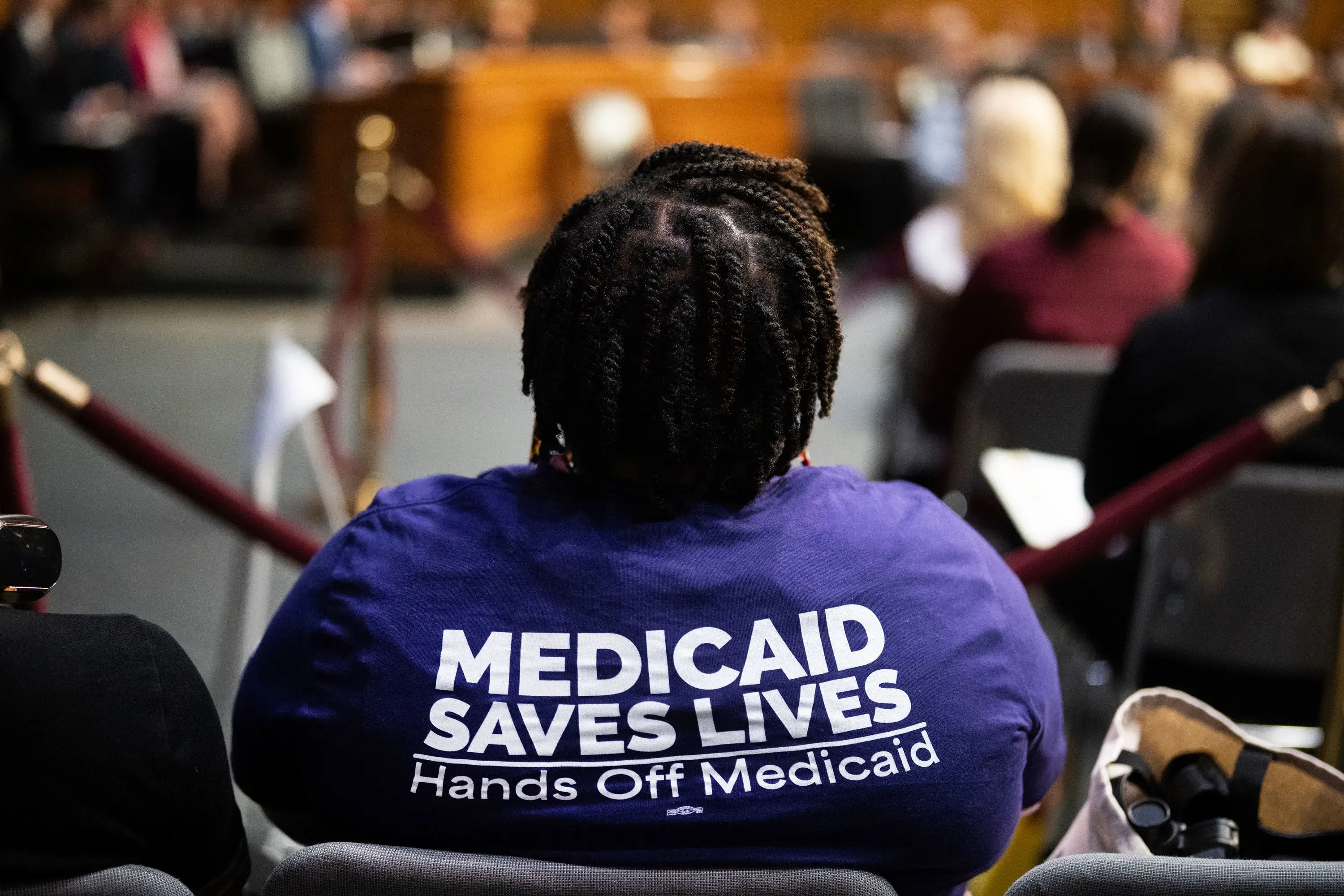 امرأة ترتدي قميصًا أرجوانيًا يحمل عبارة "Medicaid Saves Lives" خلال جلسة استماع، تعبر عن دعمها لبرنامج Medicaid.