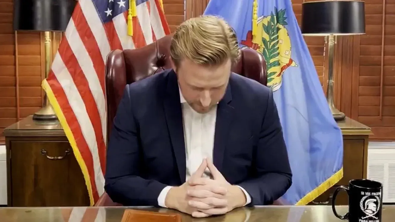 رجل جالس في مكتب، ينظر إلى الطاولة بأمامه، بينما ترفرف الأعلام الأمريكية وأوكلاهوما خلفه.