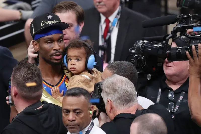 لاعب كرة سلة يحمل طفلاً يرتدي سماعات أذن، محاطاً بحشود من المعجبين والإعلام بعد فوز فريق أوكلاهوما سيتي ثاندر بلقب الدوري.