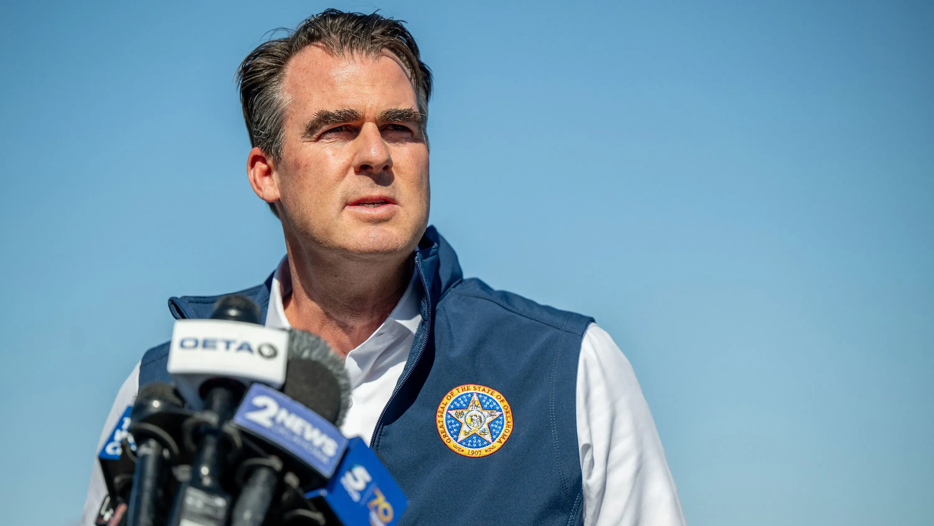 كيفن ستيت، حاكم أوكلاهوما، يتحدث في مؤتمر صحفي حول التعليم وقرارات مجلس التعليم، مع سماء زرقاء في الخلفية.