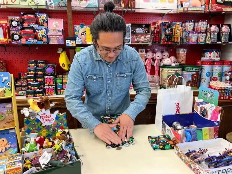 رجل يقوم بتغليف لعبة في متجر كيبز توي لاند، مع خلفية مليئة باللعب والألعاب، مما يعكس تاريخ المتجر الطويل في لوس أنجلوس.