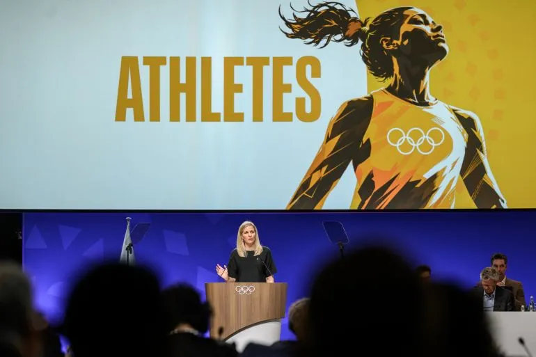 خلال اجتماع اللجنة الأولمبية الدولية، تظهر رئيسة اللجنة Kirsty Coventry وهي تتحدث عن دعم الرياضيين، مع خلفية تحمل كلمة "ATHLETES".