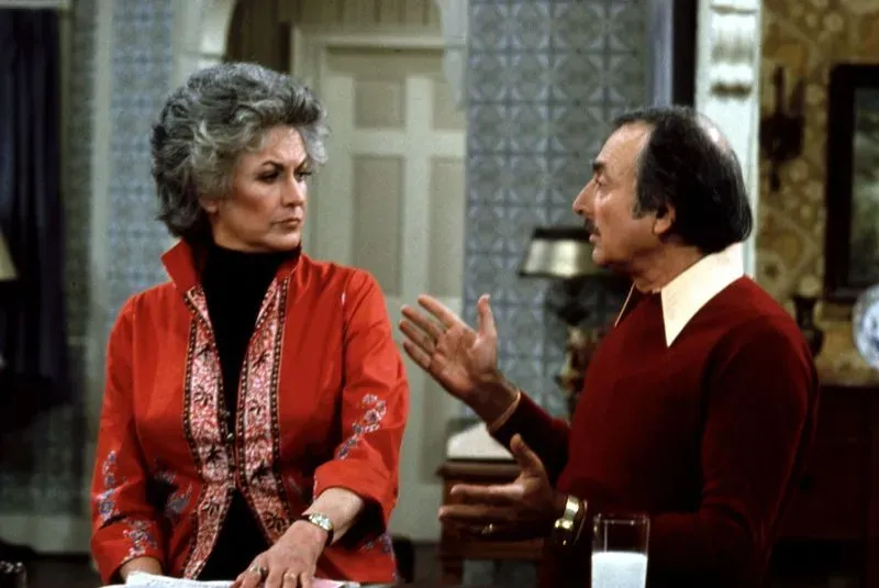 مشهد من مسلسل كوميدي يظهر شخصيتين تتبادلان الحديث بجدية، مما يعكس التوتر والسخرية في مواجهة الأزمات الاقتصادية.