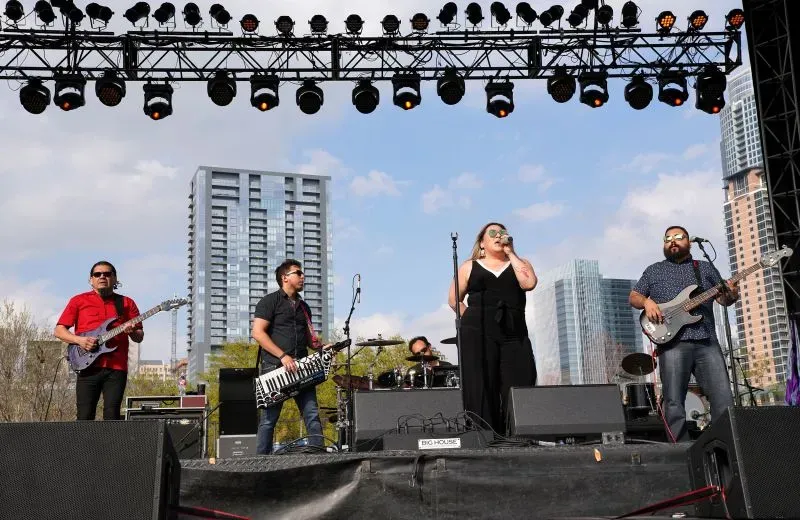 أعضاء فرقة سيلينا التكريمية يؤدون على المسرح في مهرجان ساوث باي ساوث ويست بأوستن، مع خلفية مباني حضرية.