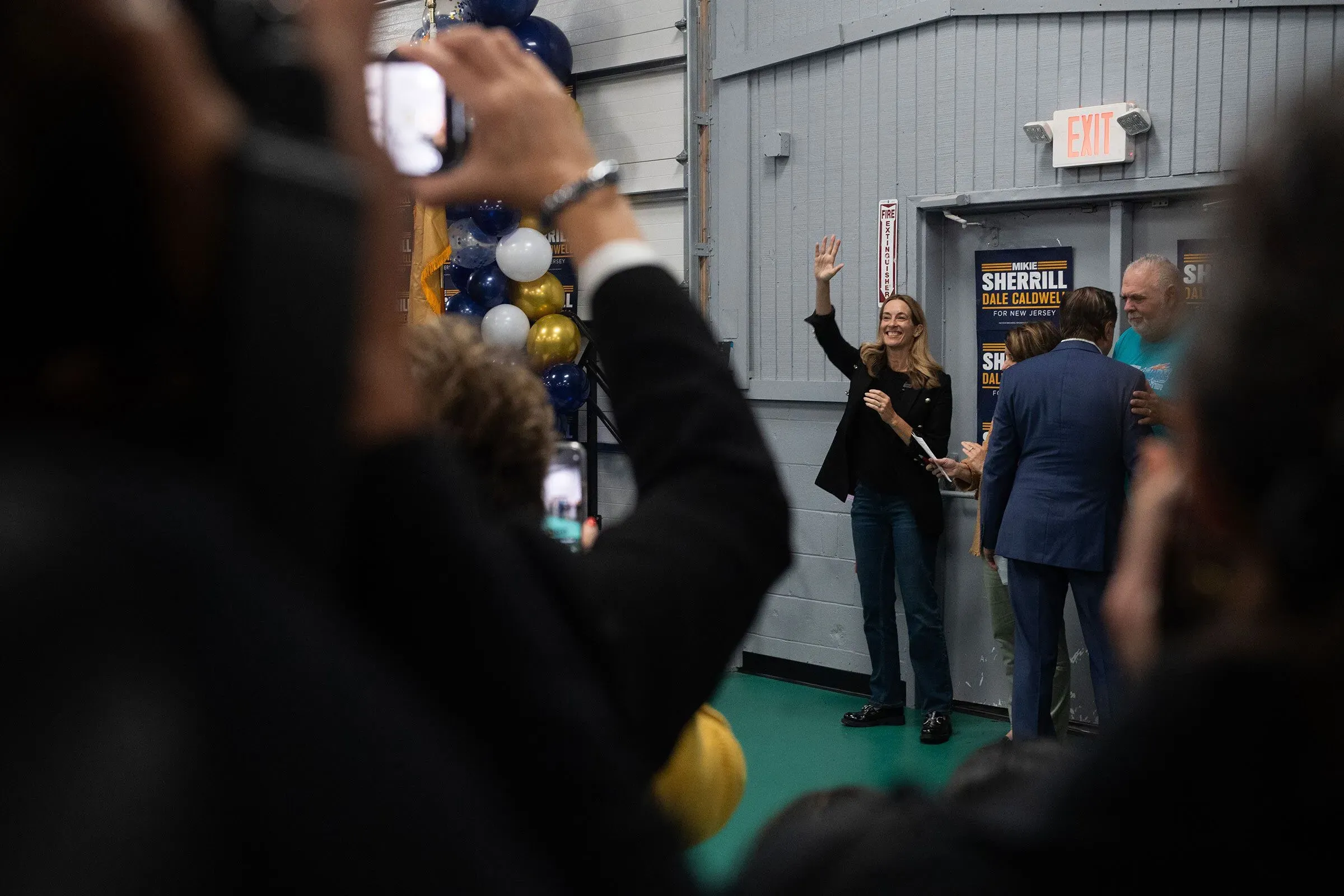 أبيغيل شيريل تلوح بيدها للجمهور في تجمع انتخابي، محاطة ببالونات زرقاء وبيضاء، تعكس أجواء الحملة الانتخابية في نيوجيرسي.