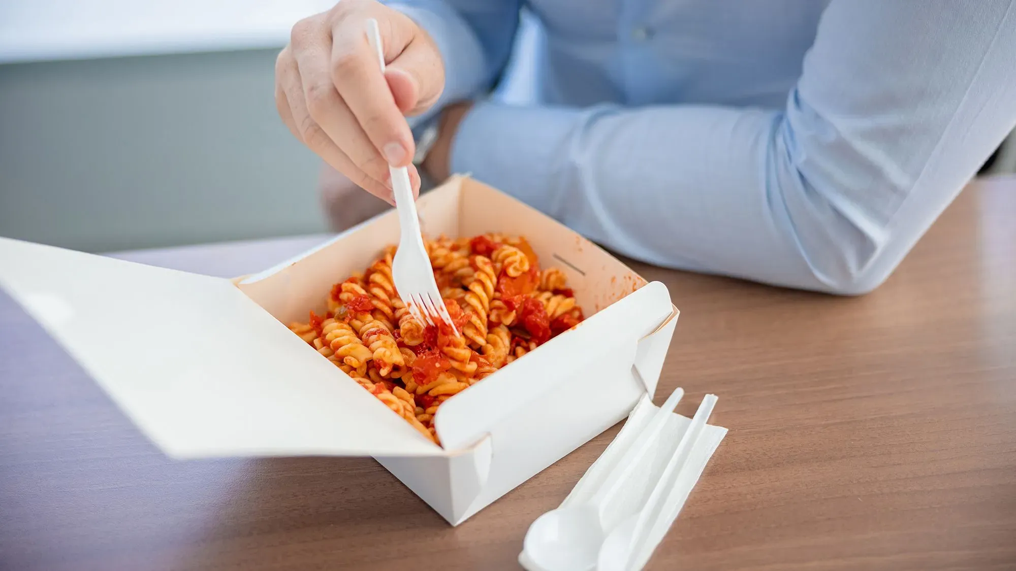شخص يستخدم شوكة بلاستيكية لتناول المعكرونة في علبة طعام، مع أدوات مائدة بلاستيكية أخرى بجانبها، مما يبرز أزمة التلوث البلاستيكي.