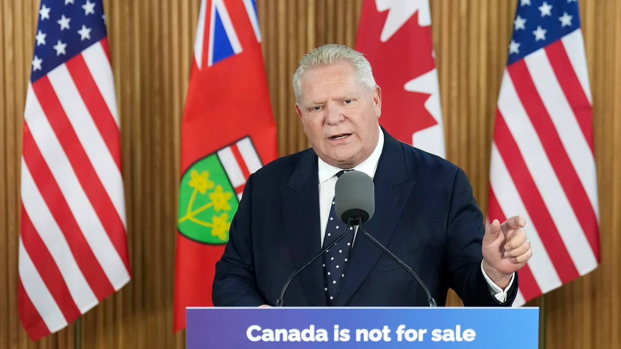 دوغ فورد، رئيس وزراء أونتاريو، يتحدث في مؤتمر صحفي أمام أعلام كندا والولايات المتحدة، مُشيرًا إلى تأثير الرسوم الجمركية على التجارة.