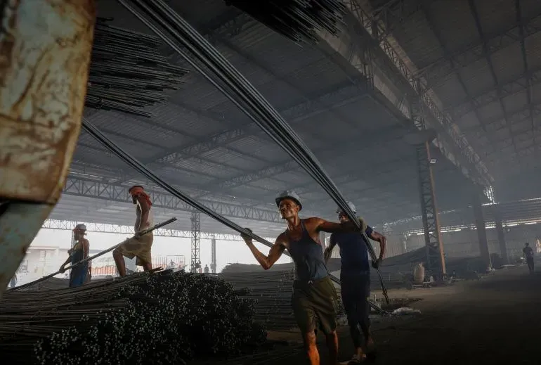 عمال يحملون قضبان الحديد في مصنع للصلب، وسط أجواء دافئة ومظلمة، مما يعكس ظروف العمل الصعبة في الصناعة الهندية.