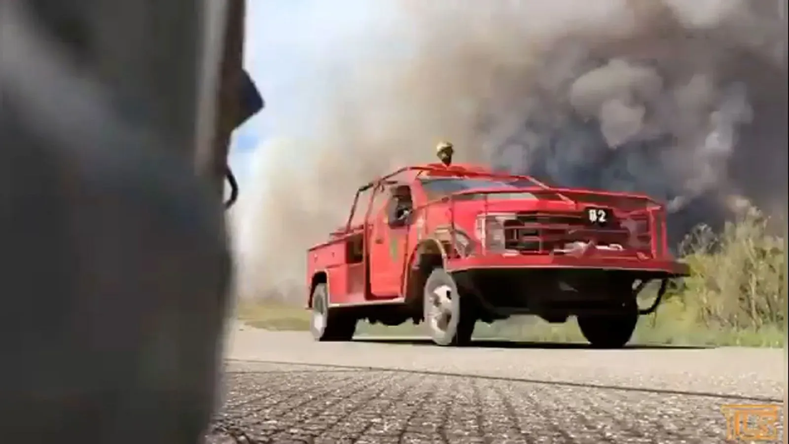 سيارة إطفاء حمراء تسير على طريق، بينما تتصاعد سحب الدخان الكثيف من حريق غابات في نيوجيرسي، مما يهدد المباني ويؤدي إلى إجلاء السكان.