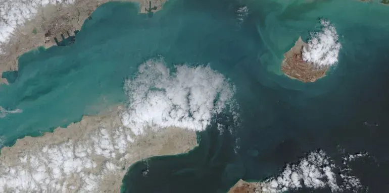 صورة فضائية لمضيق هرمز، تظهر المياه الزرقاء المحيطة بجزيرة صغيرة، مع سحب بيضاء تتناثر في السماء. تعكس الصورة التوترات الجارية بين الولايات المتحدة وإيران.