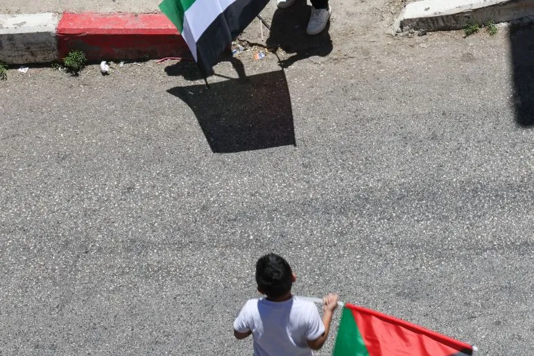 طفل فلسطيني يحمل علم فلسطين وسط شارع رملي، في مشهد يعكس التوترات والاحتجاجات المستمرة في الأراضي الفلسطينية.