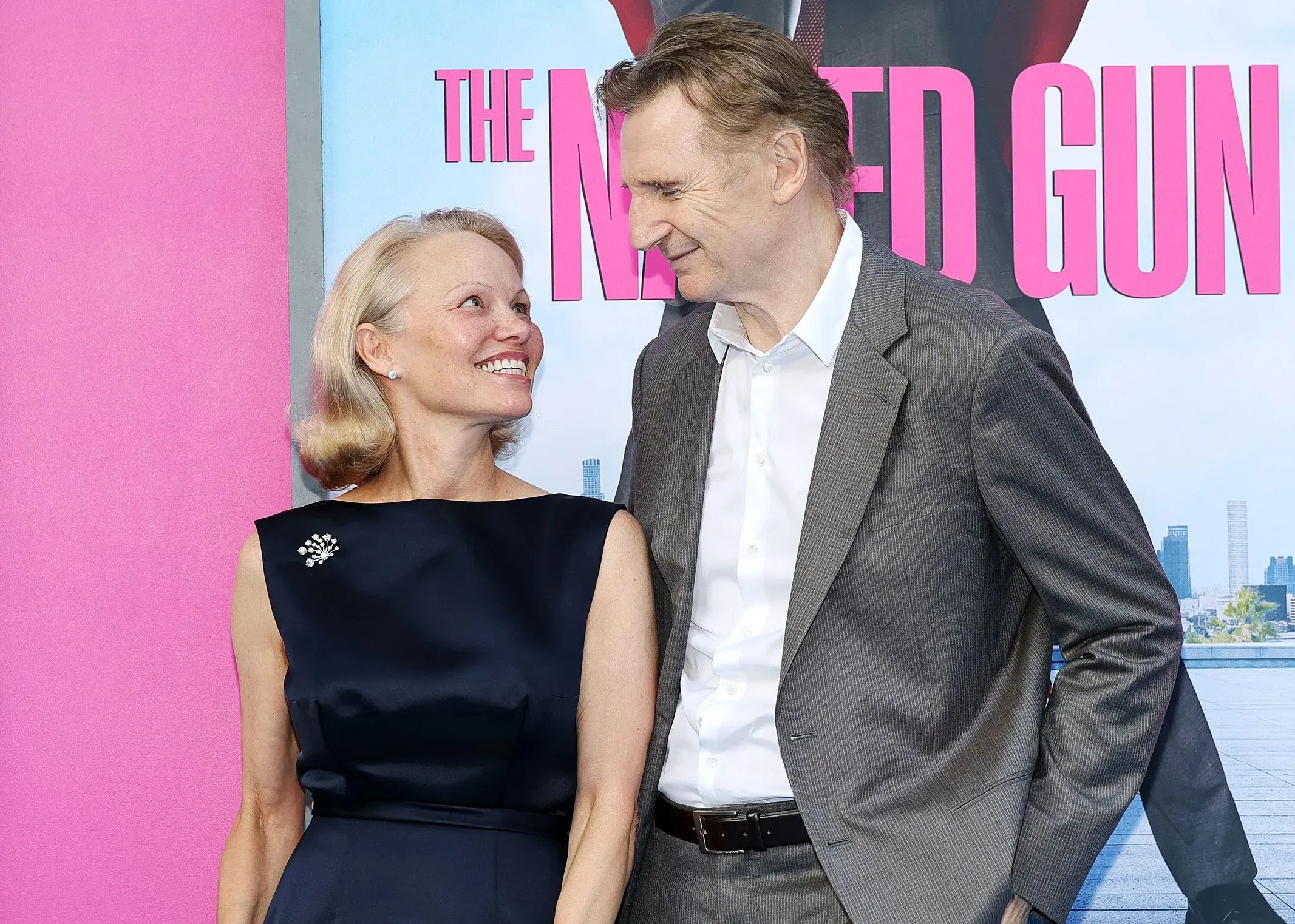 باميلا أندرسون وليام نيسون يتبادلان نظرات الحب أثناء ترويجهما لفيلم "المسدس العاري"، مع خلفية ملونة تعكس أجواء الكوميديا.