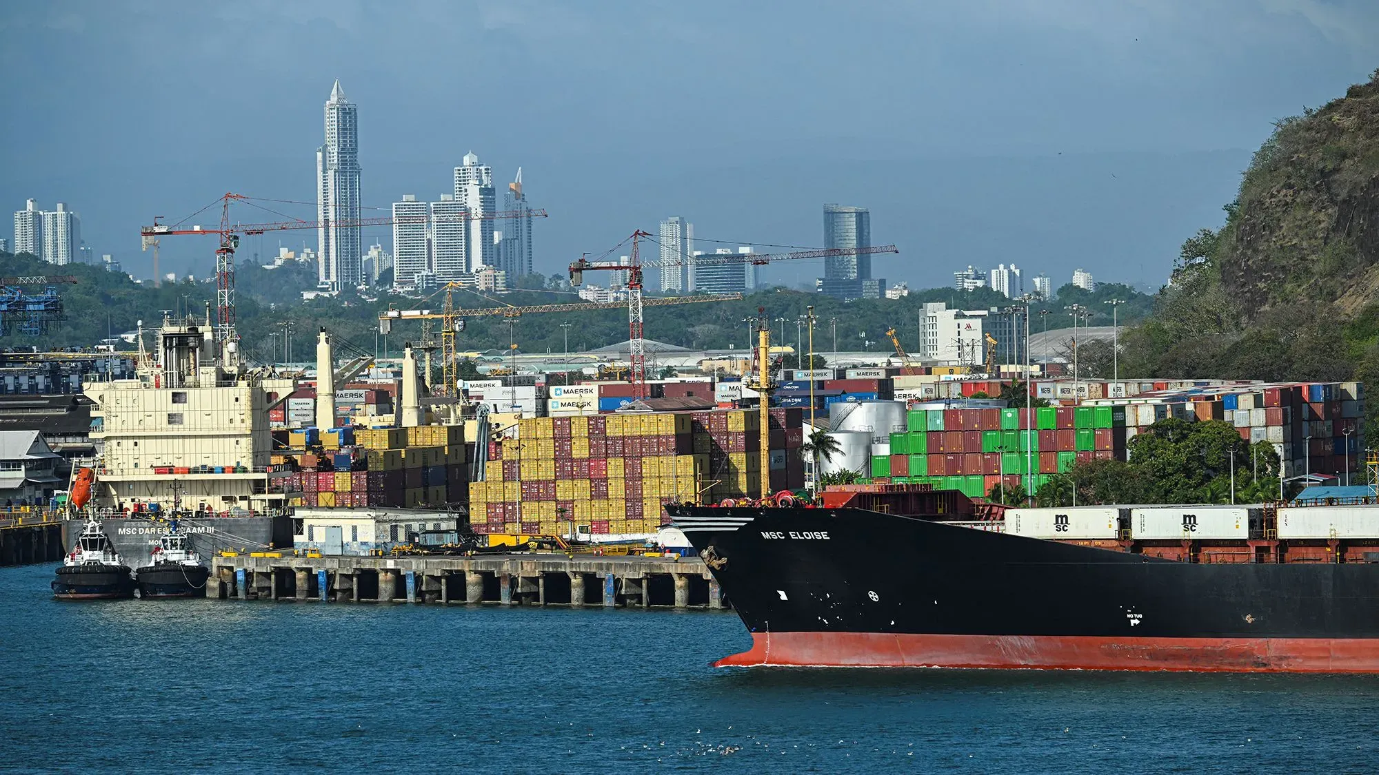 ميناء مزدحم في منطقة قناة بنما، يظهر سفينة شحن كبيرة محملة بالحاويات، مع خلفية لمدينة حديثة وأبراج.