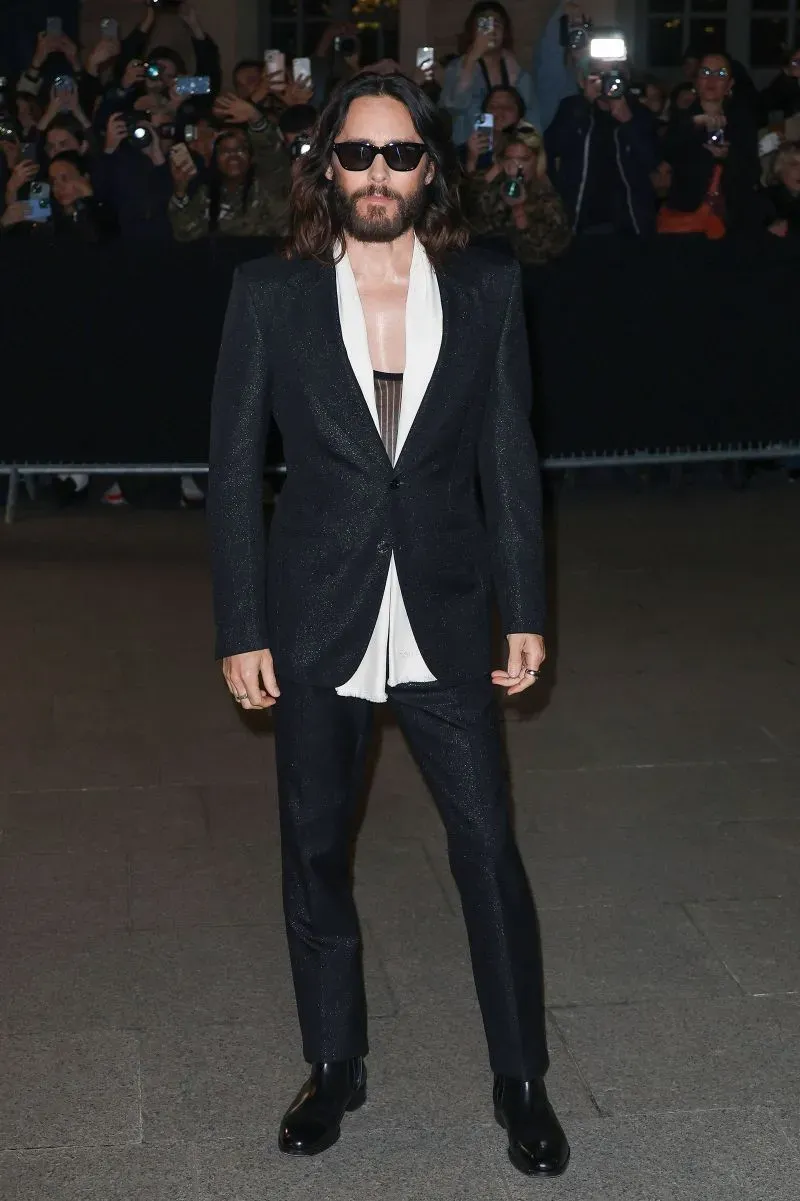 جارد ليتو يرتدي بدلة أنيقة باللون الأسود مع قميص أبيض مفتوح، بينما يتجمع المعجبون خلفه في حدث أسبوع الموضة في باريس.