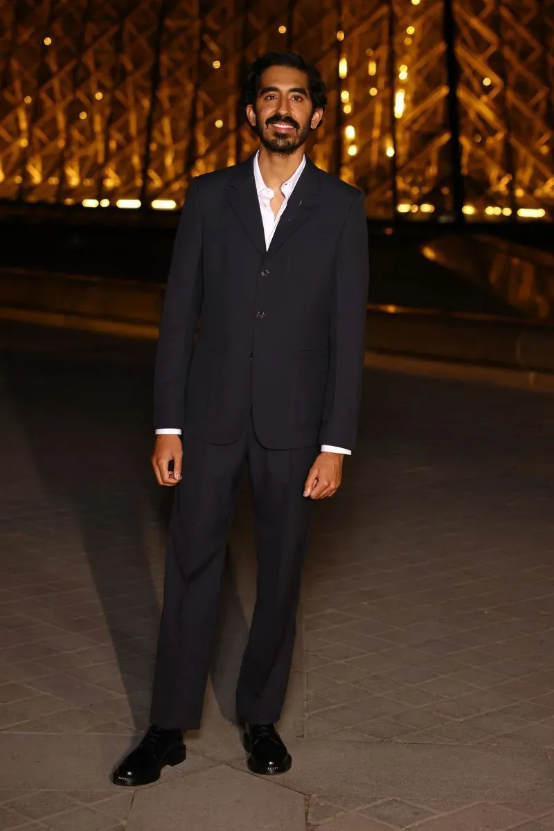 حضور شاب يرتدي بدلة أنيقة باللون الأسود، يقف أمام خلفية مضاءة بمصابيح دافئة، خلال أسبوع الموضة في باريس.