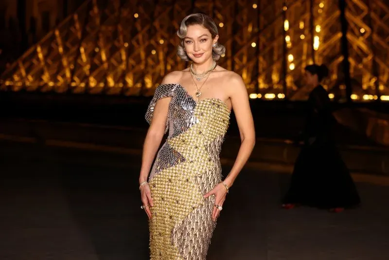 جيجي حديد ترتدي فستاناً مميزاً بتصميم لامع أمام متحف اللوفر خلال أسبوع الموضة في باريس، مع إضاءة خلفية ساحرة.
