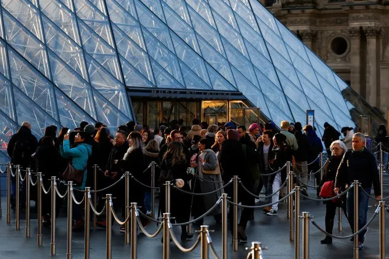 طابور من الزوار عند مدخل الهرم الزجاجي لمتحف اللوفر، مع الإضاءة الساطعة خلفهم، في ظل مساعي الترميم للحفاظ على الأعمال الفنية.