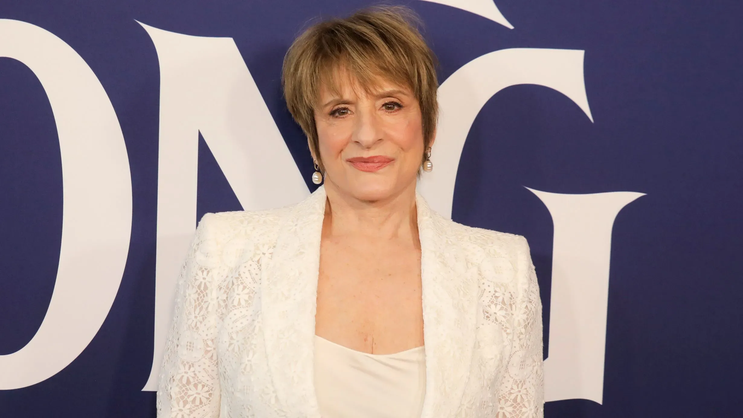 باتي لوبون ترتدي بدلة بيضاء وتبتسم في حدث برودواي، وسط خلفية تحمل شعار "تون". تعكس الصورة الجدل المحيط بتصريحاتها الأخيرة.