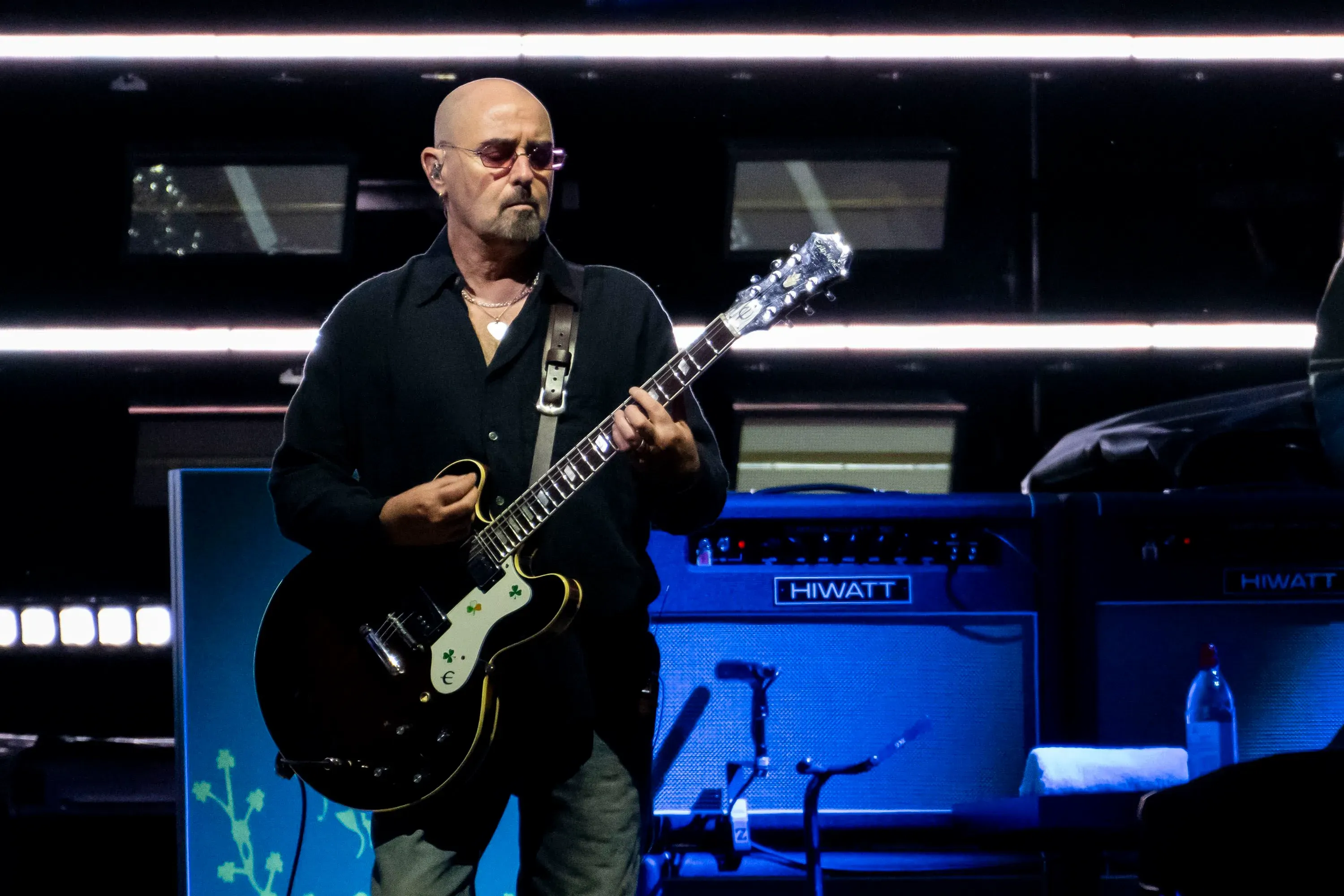 بول آرثرز، عازف الجيتار في فرقة Oasis، يؤدي على المسرح، مع وجود أمبيرات موسيقية خلفه، أثناء جولة الفرقة.