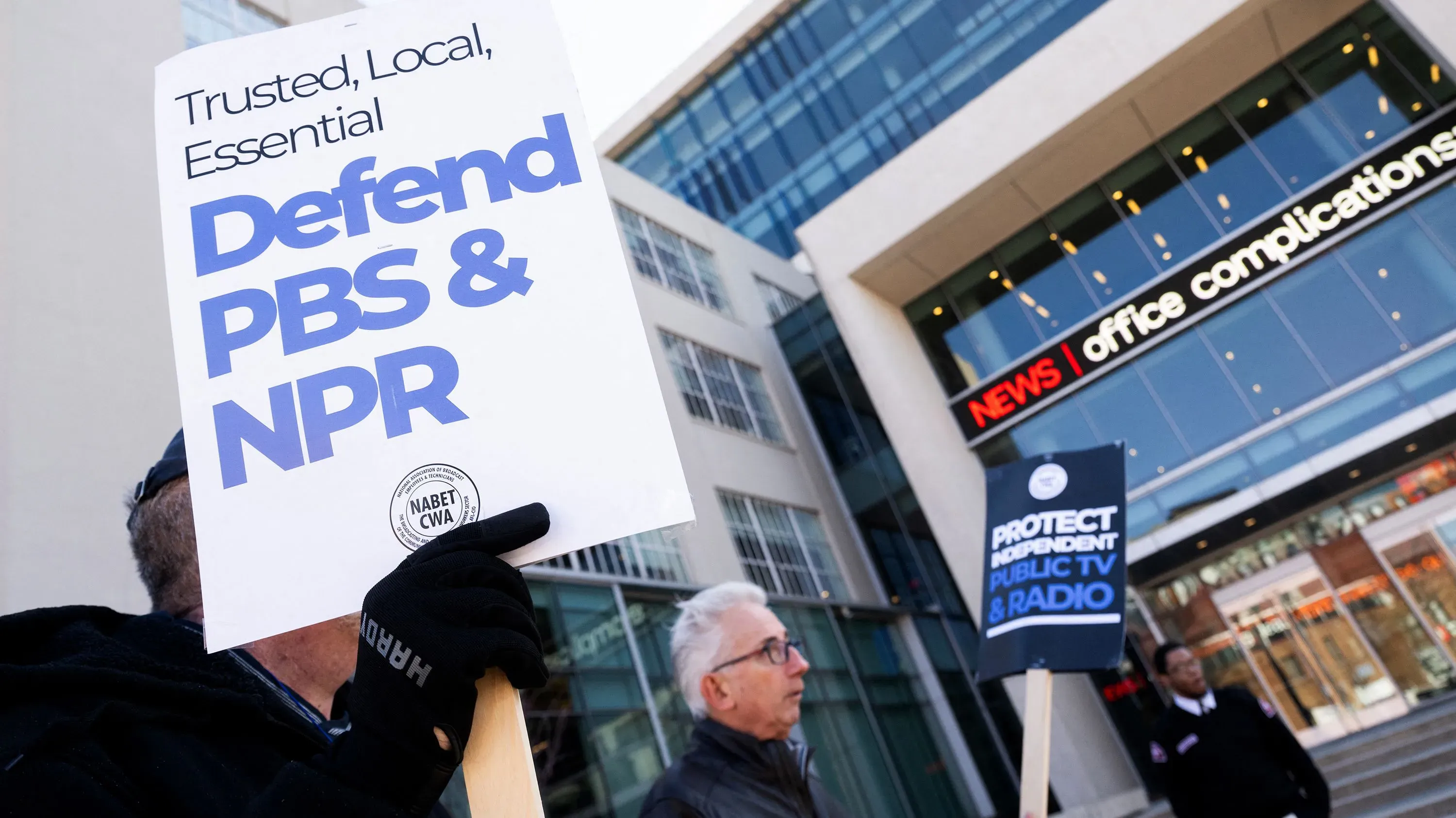 لافتات تحمل شعارات تدعو للدفاع عن PBS وNPR، مع متظاهرين أمام مبنى إخباري، تعكس الجهود لحماية التمويل العام للإعلام.