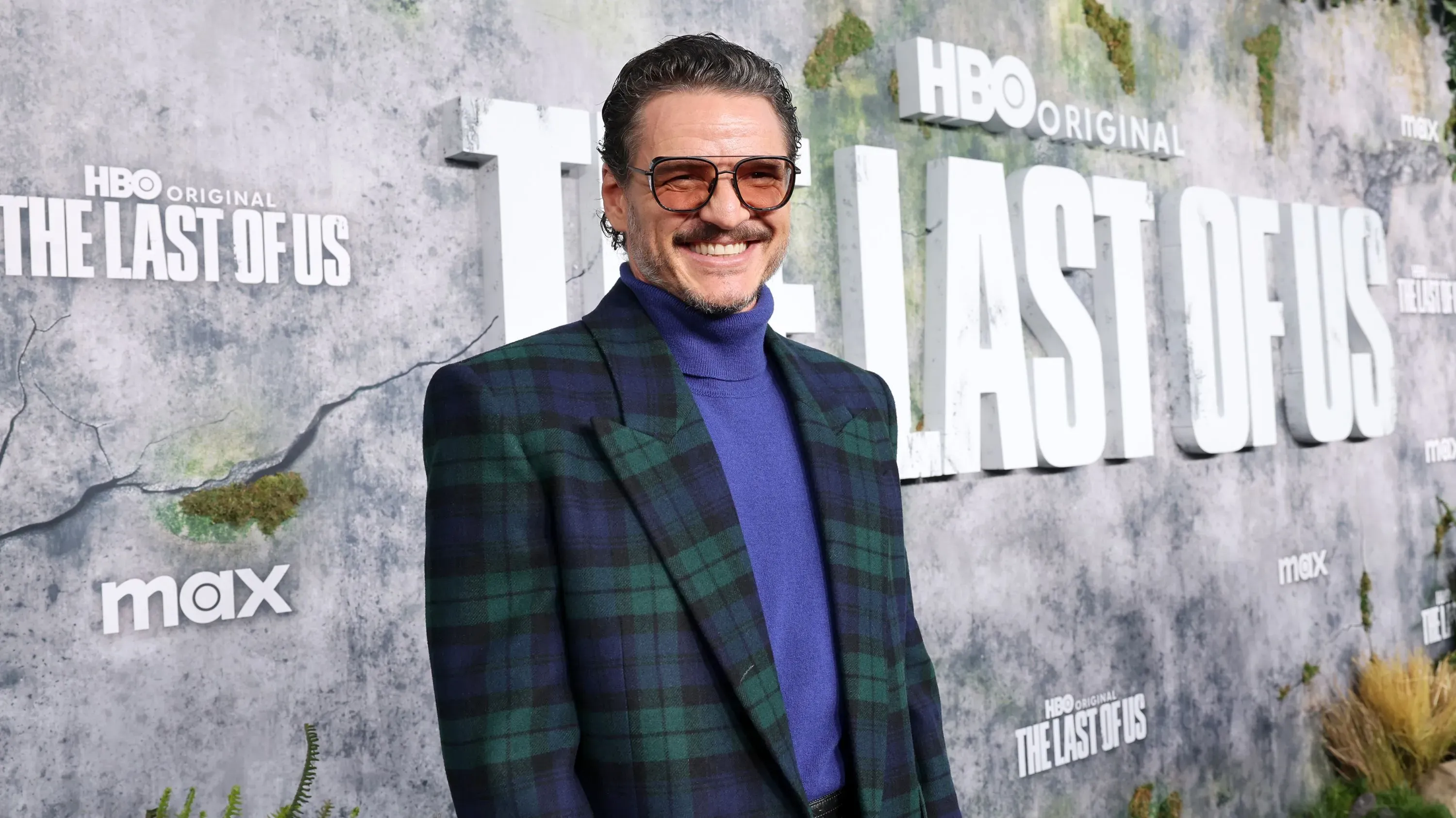 باسكال يرتدي ملابس أنيقة ويبتسم في عرض مسلسل "آخرنا" على HBO، مع خلفية تحمل اسم المسلسل.