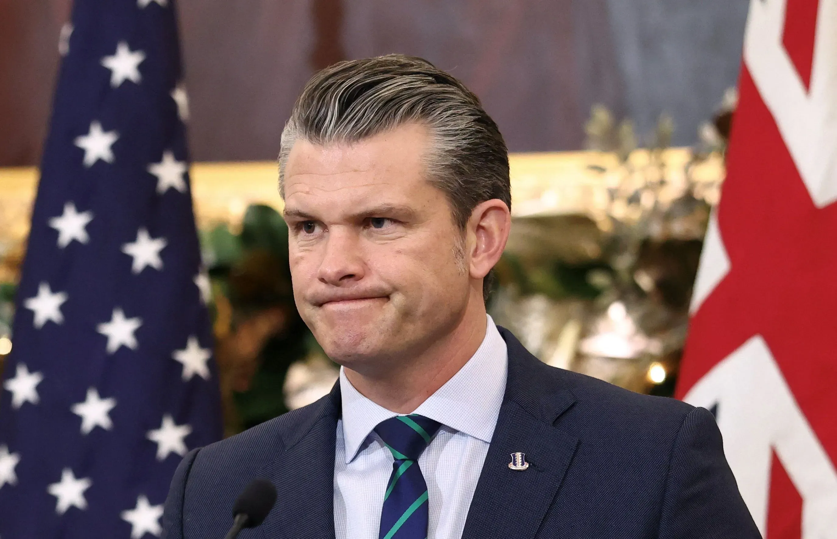 وزير الدفاع بيت هيغسيث يظهر بتعبير جاد، محاطًا بعلم الولايات المتحدة وعلم آخر، في سياق حديث حول تسريبات معلومات حساسة.