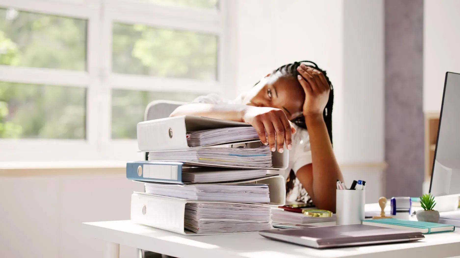 امرأة شابة تعاني من التوتر والإرهاق الجسدي، تتكئ على مكتب مليء بالمجلدات والملفات، تعكس ضغوط الحياة اليومية وتأثير اضطراب نقص الانتباه وفرط الحركة.