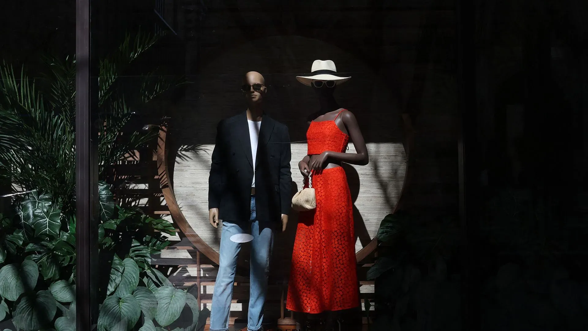 عرض لتمثالين لعارضات أزياء في واجهة متجر، حيث ترتدي إحداهن فستانًا أحمر وتظهر الأخرى ببدلة عصرية، مع خلفية نباتات خضراء.