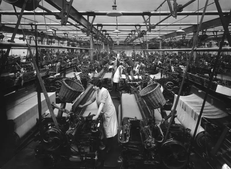 نساء يعملن في مصنع للملابس، حيث تظهر آلات النسيج والعملية الإنتاجية، مما يعكس تأثير التعريفات الجمركية على صناعة الملابس.