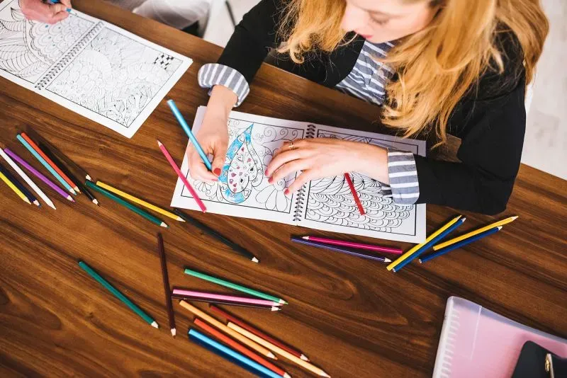 امرأة شابة تلوّن رسومات في دفتر على طاولة، محاطة بأقلام تلوين ملونة، تعبر عن الإبداع والعودة إلى الهوايات القديمة.