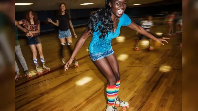 امرأة تتزلج بسعادة على الزلاجات في حلبة، مرتدية قميصًا أزرق وسروال قصير، مع أصدقاء حولها، تعكس لحظات من المرح والحنين إلى الطفولة.