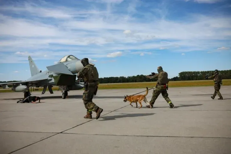 عناصر من الجيش البولندي يقومون بعمليات أمنية على مدرج مطار، مع وجود طائرة مقاتلة في الخلفية، في إطار تعزيز الدفاعات الجوية.
