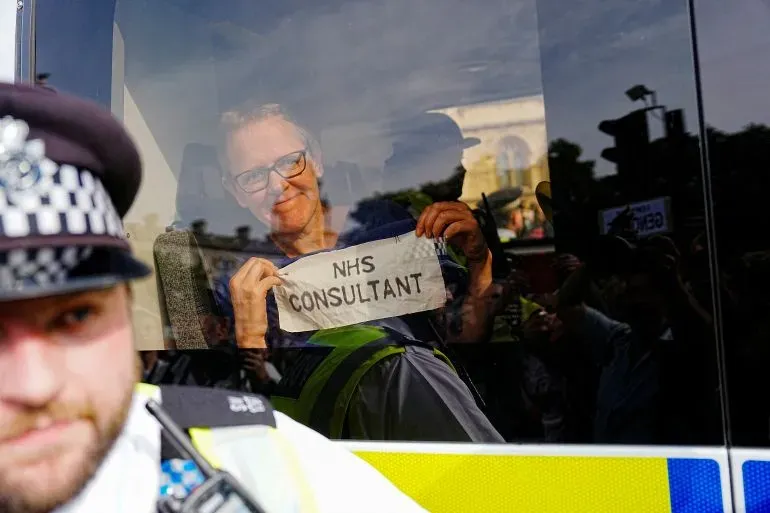 رجل مبتسم يحمل لافتة مكتوب عليها "استشاري NHS" داخل سيارة شرطة، بينما يظهر ضابط شرطة في المقدمة خلال مظاهرة في لندن.