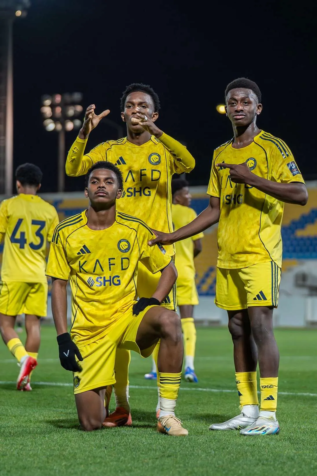ثلاثة لاعبين شباب من فريق النصر السعودي يرتدون الزي الأصفر يحتفلون في ملعب مضاء استعدادًا لمباريات دوري Joy Elite تحت 21 سنة.