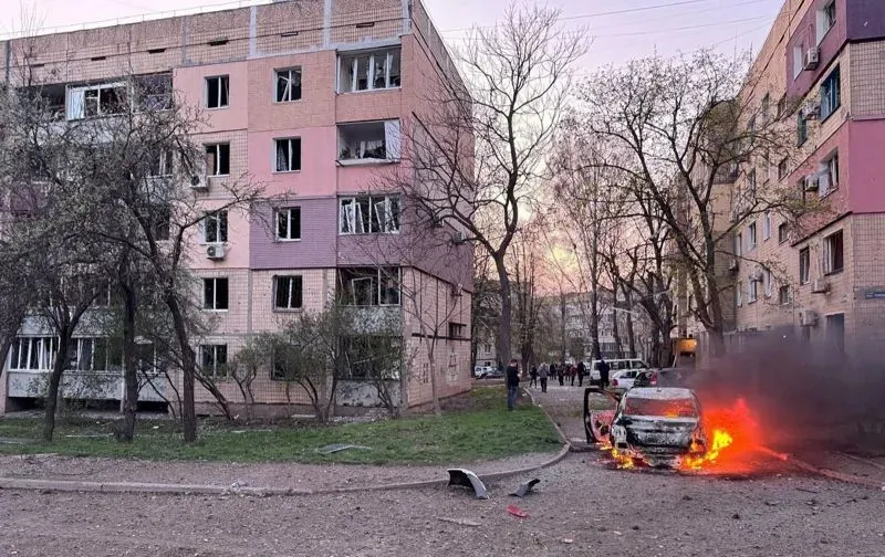 مبنى سكني متضرر في أوكرانيا، مع سيارة مشتعلة في المقدمة، مما يعكس آثار الحرب والدمار في المنطقة.