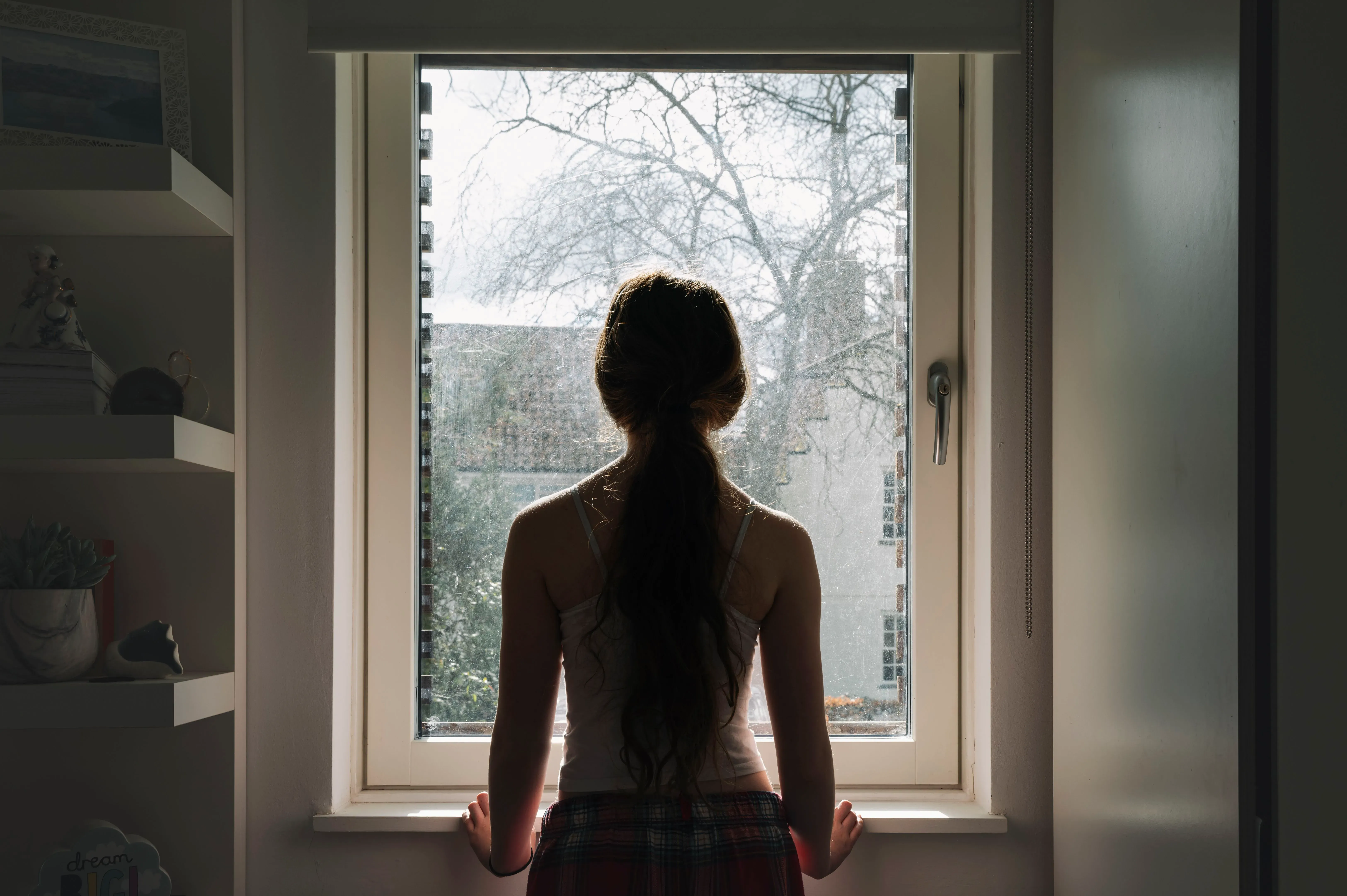 فتاة صغيرة تقف أمام نافذة، تنظر إلى الخارج في ضوء النهار، تعكس مشاعر العزلة والقلق المرتبطة بزيادة حالات إيذاء النفس بين الأطفال.